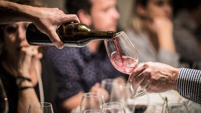 A glass of red wine is poured. In the background people are eating and smiling.