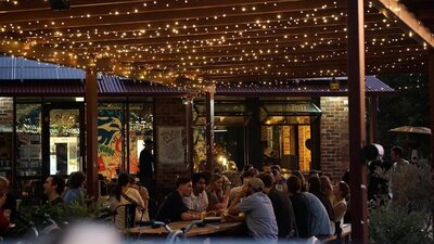 People sitting along outdoor tables in the evening time, underneath fairy lights