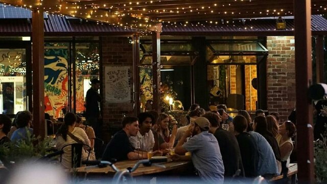 People sitting along outdoor tables in the evening time, underneath fairy lights