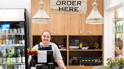 A woman behind a cafe counter underneath a sign that says 'order here',  ready to serve customers
