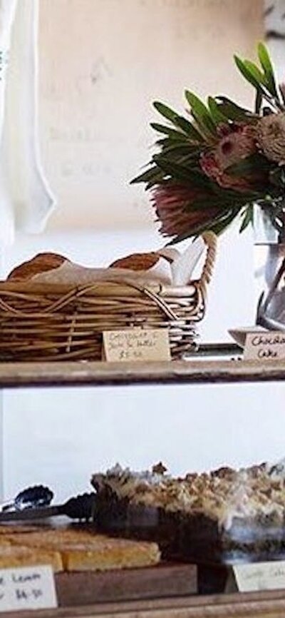 Cake cabinet full of homemade cakes. Fresh flowers sit on the bench.