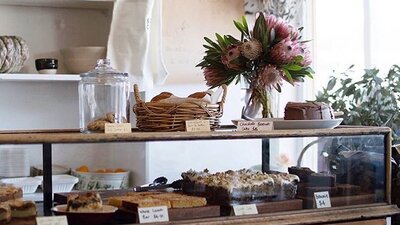 Cake cabinet full of homemade cakes. Fresh flowers sit on the bench.