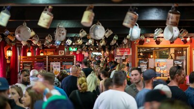 Crowd of people inside a pub, with different bottles and vessels hanging form the ceiling