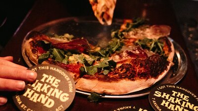 People grabbing at a pizza. The Shaking Hand branded coasters surrounding pizza