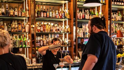 Bartender pouring cocktail into glass at bar with people waiting at bar