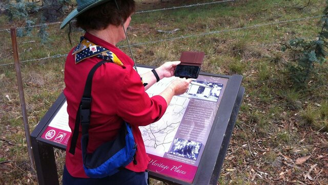 Get more from your visit with the CBR Tracks App triggering 2 min videos Woman holding smart phone in front of image on sign located in the bush