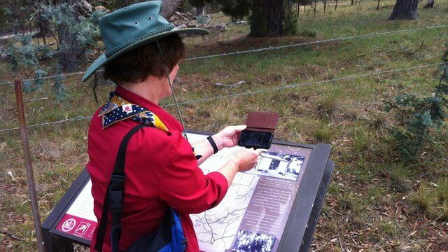 Get more from your visit with the CBR Tracks App triggering 2 min videos Woman holding smart phone in front of image on sign located in the bush