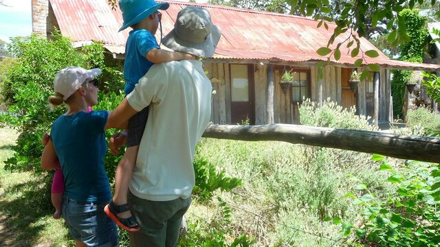 An old slab hut in the middle of suburbia has a story to tell Young family in summer clothes and hats looking at an old wooden hut