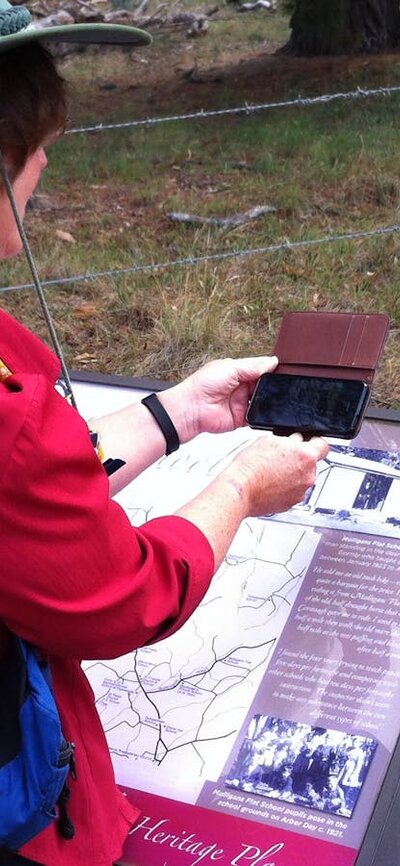 Woman in red shirt holding smart phone over an information panel in bush