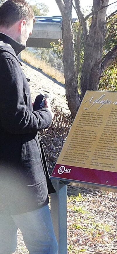 Man reading a sign board in treed area with road bridge in background