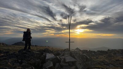 A hiker standing at the summit of a mountain with the sun setting behind them.