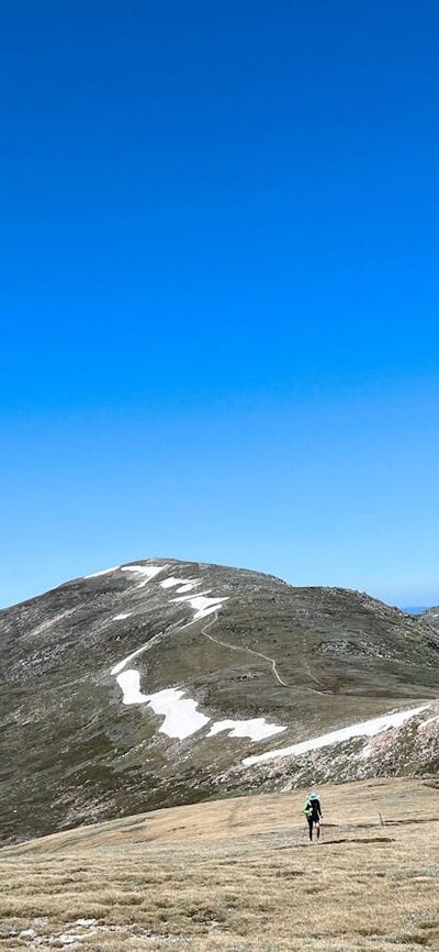 A hiker in the mountains climbing up a steady hill with snow in the background.