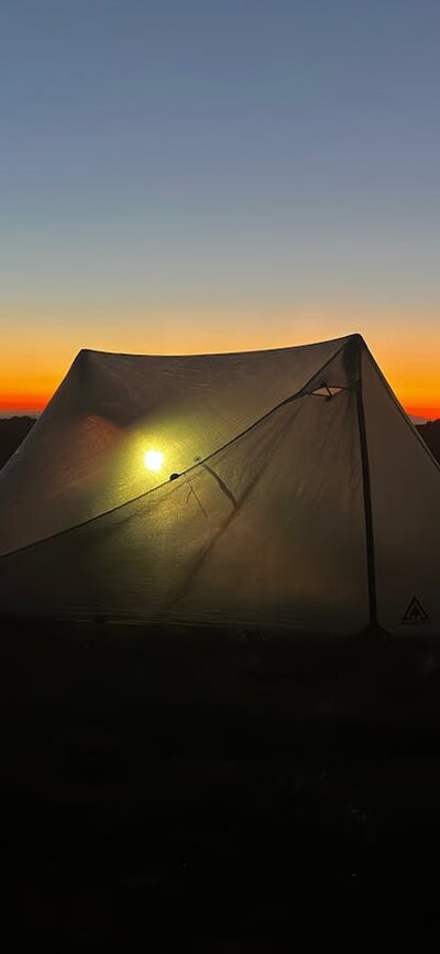 A tent in the mountains with the sun setting behind it.