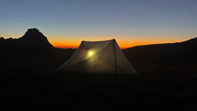 A tent in the mountains with the sun setting behind it.