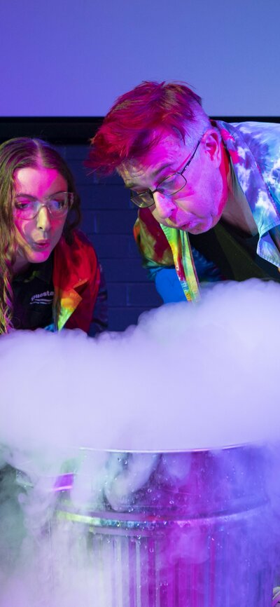 Image of two adult scientists in tye-dye lab coats that are glowing, blowing smoke out of a science experiment at Questacon. | © Martin Ollman