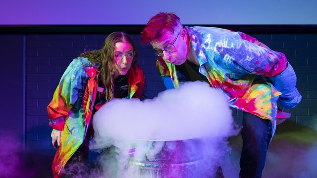 © Martin Ollman Image of two adult scientists in tye-dye lab coats that are glowing, blowing smoke out of a science experiment at Questacon. | © Martin Ollman