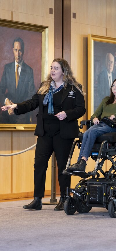 Young female tour guide shows a young woman in a mobility chair through portraits hung on the walls inside Australian Parliament House.