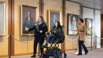 Accessible tours through Australian Parliament House Young female tour guide shows a young woman in a mobility chair through portraits hung on the walls inside Australian Parliament House.