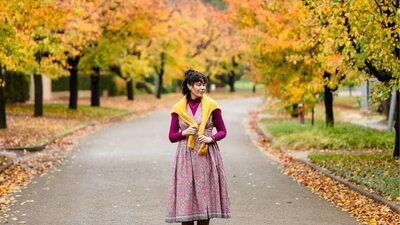 © @welch Image of a woman in a pink dress with yellow sweater standing in the middle of an autumn-leave-lined street. | © @welch