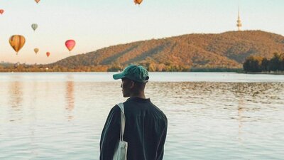 © @jefordmillare Image of a man in a ball cap standing in front of Lake Burley Griffin with hot air balloons across the sky | © @jefordmillare
