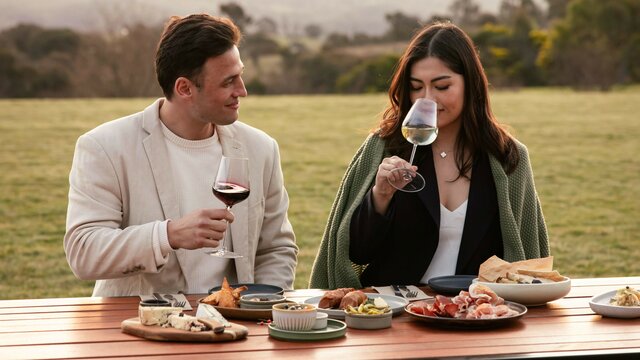 Two patrons sitting outside at a cellar door with wine and nibbles on a table.