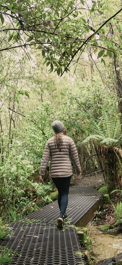 Image of a woman in a light puffer jacket and hat walking the Cascade Trail through trees in Tidbinbilla Nature Reserve. | © We Are Explorers