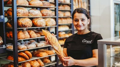 A person holds a loaf of bread in front of a diverse array of baked goods behind them.