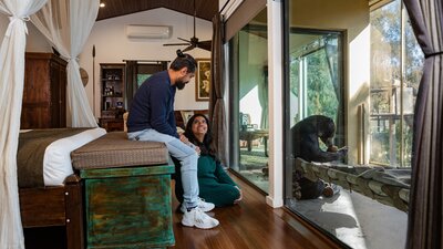 A couple looking at each other in a luxury hotel room that looks out upon a sun bear eating food.