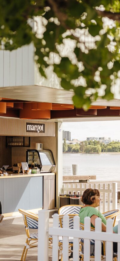 A vibrant café/bar located by Lake Burley Griffin. The outdoor seating area features modern tables and chairs with colourful cushions, surrounded by lush greenery. 