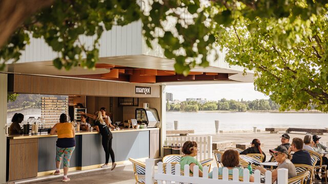 A vibrant café/bar located by Lake Burley Griffin. The outdoor seating area features modern tables and chairs with colourful cushions, surrounded by lush greenery. 
