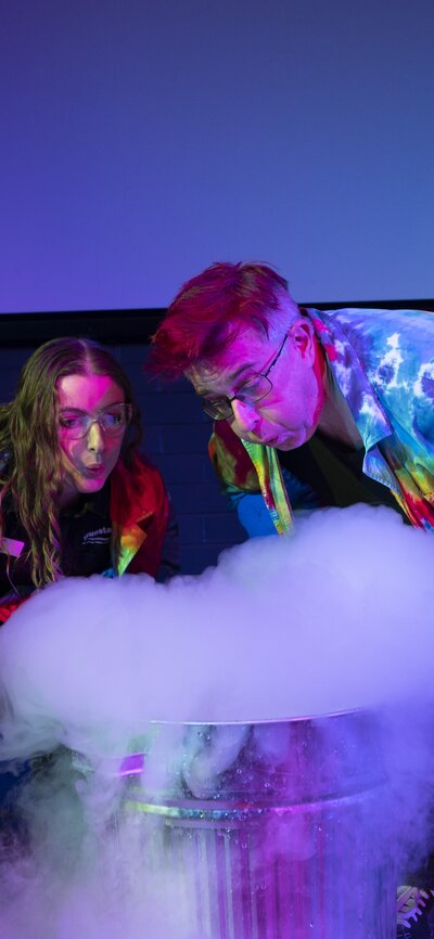 Two people wearing rainbow coloured tie dye lab coats, standing behind a liquid nitrogen science experiment. 
