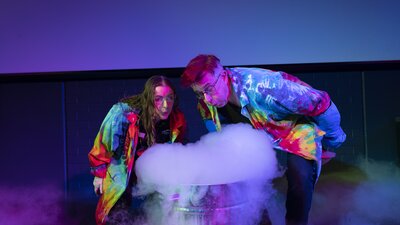 Two people wearing rainbow coloured tie dye lab coats, standing behind a liquid nitrogen science experiment.