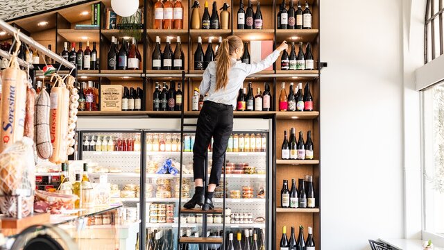 A woman climbing a ladder to a tall shelf of deli food