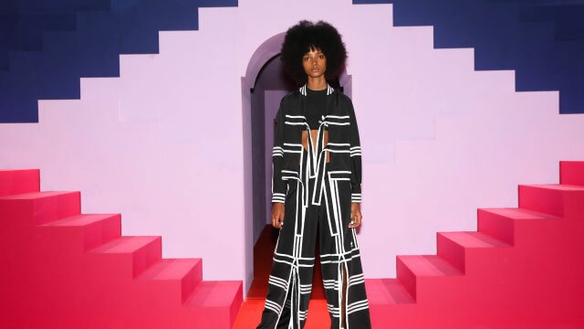 Person in a black and white patterned outfit stands on a red carpet in front of a vibrant geometric backdrop with pink and purple stairs and a central archway.