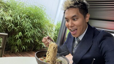 Person sitting at an outdoor table with green bamboo plants in the background, holding a forkful of noodles above a large bowl of noodle soup. The person is wearing a dark suit jacket and patterned tie.