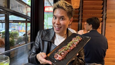 Person seated indoors at a wooden table holding a rectangular black plate with sliced grilled meat garnished with green herbs
