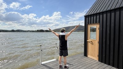 Person standing on a wooden deck beside a small black cabin with a wooden door, facing a large lake with arms raised. The scene includes calm water, distant hills, and a partly cloudy blue sky
