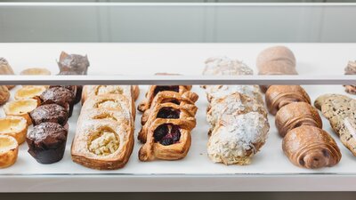 A display case filled with assorted pastries, including muffins, custard tarts, fruit danishes, almond croissants, pain au chocolat, and rustic loaves, arranged neatly on a white counter in a bakery