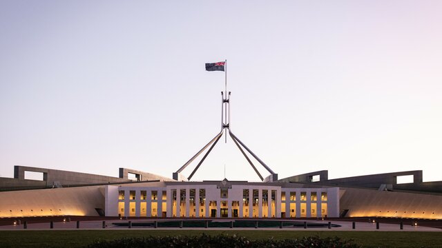 A parliament building exterior holding the Australian flag with a backdrop of a sky at sunrise