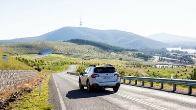 A car drives along a scenic road through Canberra’s National Arboretum, with rolling hills and distant mountains under a clear sky