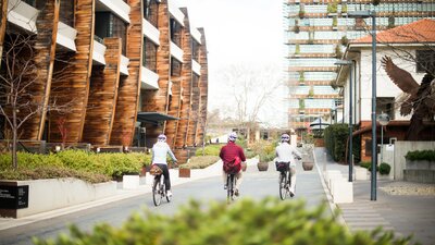 Three people cycling through the NewActon precinct in Canberra, surrounded by modern timber and glass buildings