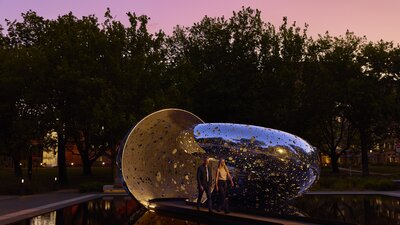 A couple walking out of a large sculpture at dusk time