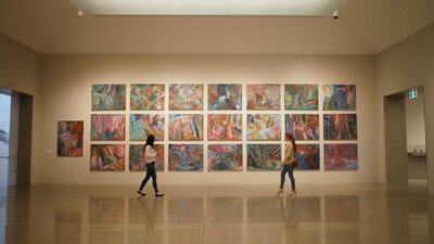 Two women walk across a wall of colourful paintings at the National Gallery of Australia
