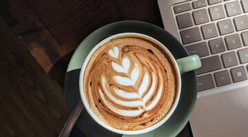 A latte in a green mug on a table with a laptop in the background