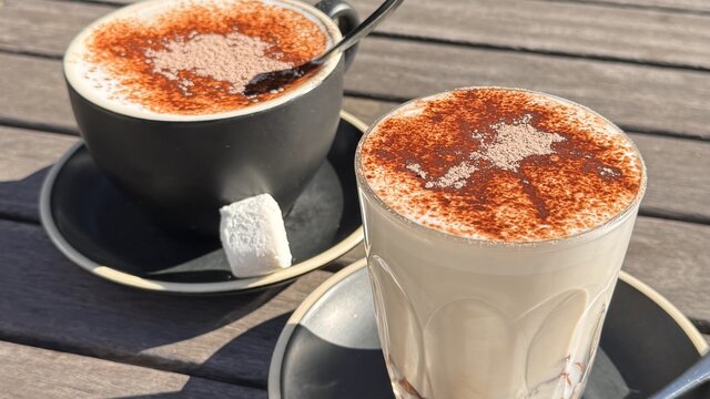 A hot chocolate in a mug and a hot chocolate in a cup, both with marshmallows and a spoon on the side