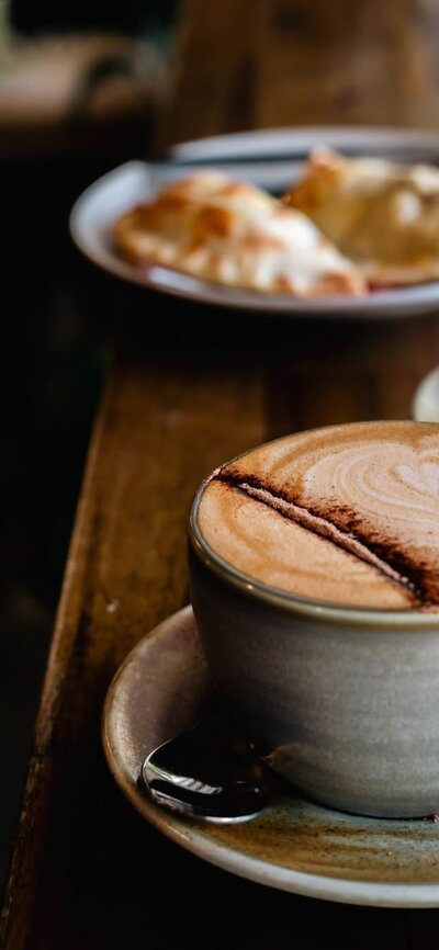 Two hot drinks in mugs with food plated in the background