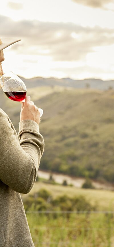 Man in a hat and grey jacket sips red wine while looking at the Brindabella Hills.