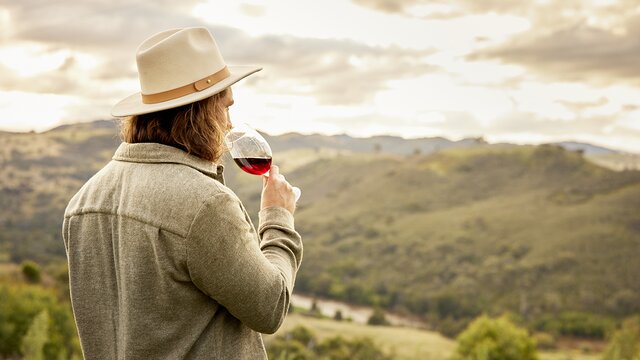 Man in a hat and grey jacket sips red wine while looking at the Brindabella Hills.