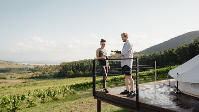 A couple pouring wine on a deck in front of a vineyard.