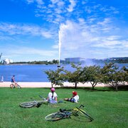 Two people with bikes relaxing on the grass in front of the lake and a jet of water.